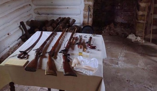 Long muskets, a sword, pistols, and cleaning equipment sits of a table in a room of the Fort Gibson Stockade with a fireplace.
