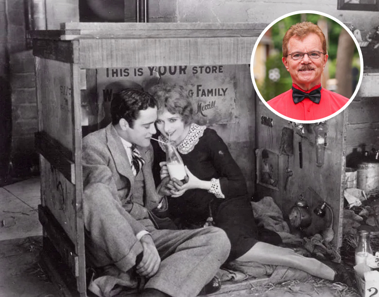 A man and a woman huddle together in a wooden box crate sharing a milk bottle, drinking through a straw.