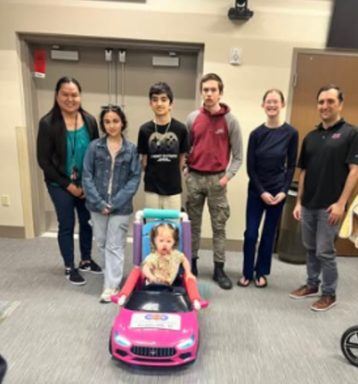 A team of six high school students and teachers stand behind a young girl in a pink toy car they adapted with a custom seat.