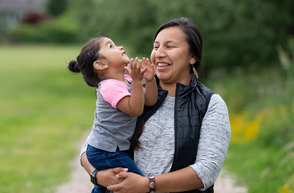 A Native American mom holds her toddler-age daughter affectionately while they take a break from walking in the park to smile at each other.