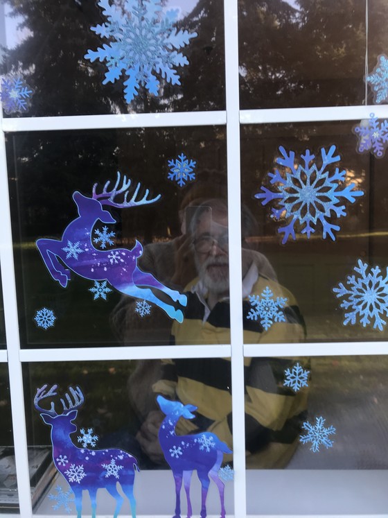 man white hair beard slightly smiling looking out window decorated with reindeers snowflakes woman taking photo with phone reflected same window