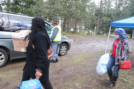 Two woman carry supplies while a person stands at minivan wearing yellow vest, face shield and mask administering test.