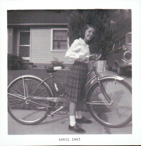 Photo of Jane standing over her bike wearing a white blouse and plaid skirt in 1963. 