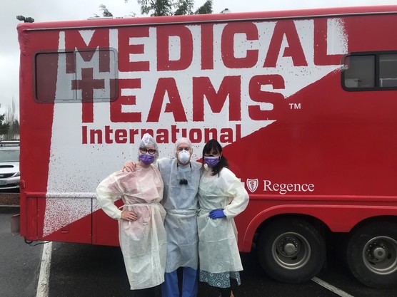 Three people in masks and PPE posing together in from of a red van that says, "Medical Teams International".