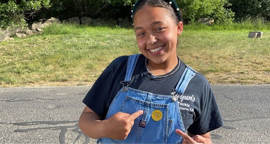 Young person smiling and pointing to the sticker on her overalls that says "I got my COVID-19 vaccine".
