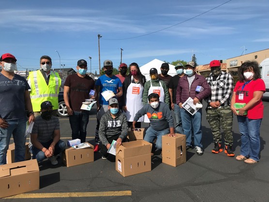 15 people wearing masks pose in a parking lot. Some hold paperwork and some are kneeling with cardboard boxes.