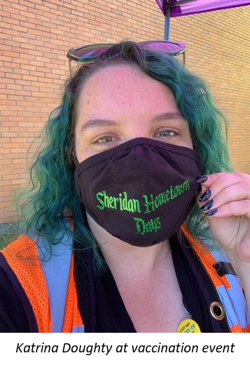 Woman with green hair, sunglasses on top of head, orange safety vest, and black mask with green gothic test saying Sheridan Hometown Days