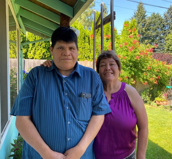 Tall man, black hair, blue shirt stands next to short woman in magenta shirt, greying dark hair who has arm around his shoulder next to a house