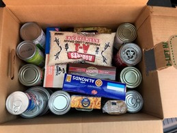Cardboard box with many cans, boxes of macaroni and cheese, almonds and mashed potatoes, a bag of rice and oatmeal.