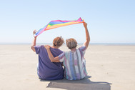 an older couple sits on the beach while holding a Pride flag