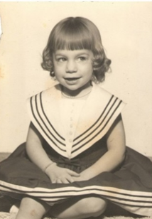 A sepia-toned childhood photo of Laurie Theodorou with bangs, ringlets and a sailor dress