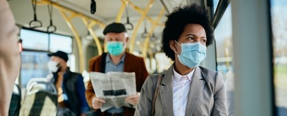 Photo: black woman wears a mask on a bus. Older man in a black hat behind standing her is in a mask and reading newspaper. 