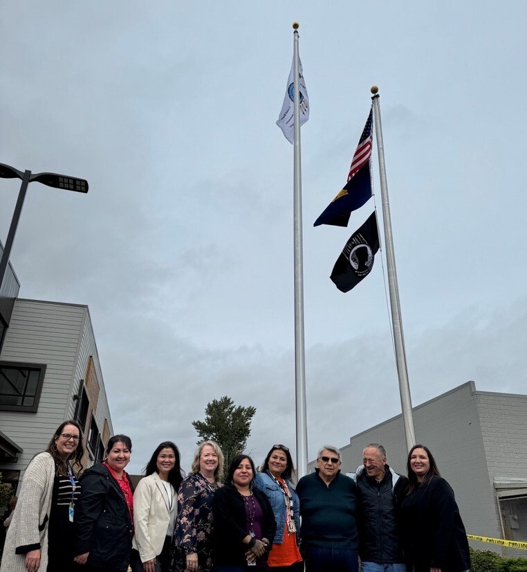 a group of people stand in front of a flag pole with the US flag, the Oregon state flag, a POW MIA flag, and the Grand Ronde Tribal flag