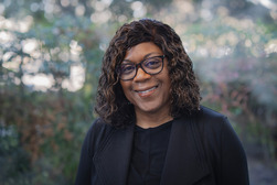 Black woman with dark shoulder length hair, wearing black and smiling in front of a green bush