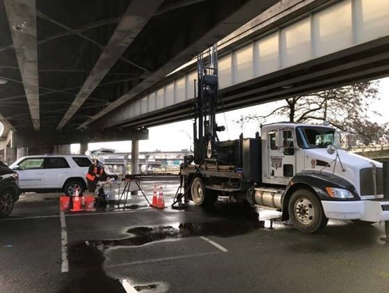 People working next to vehicles and drilling equipment in a paved area underneath a bridge.