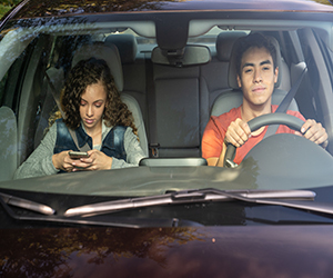 Two teens in a car - one driving and the other teen looking at her phone.
