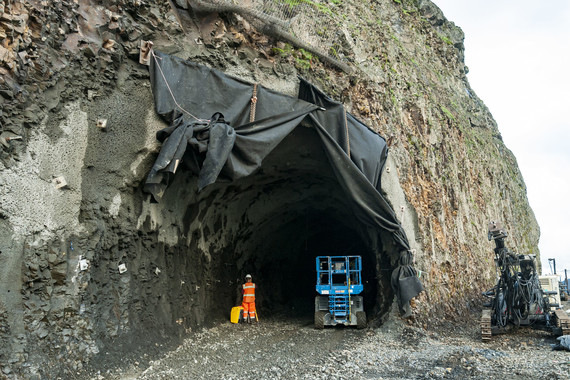 A large arched tunnel entrance in a cliff face with a crew member in reflective gearing facing into the tunnel next to lift equipment.