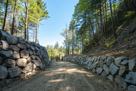 Hand stacked rock walls on either side of a wide dirt trail. Trees, blue sky and people in the distance. 