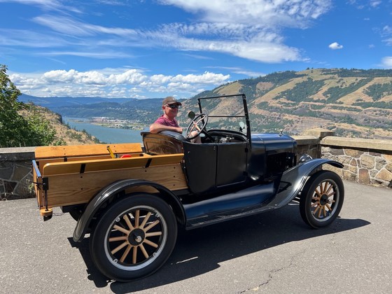 A man sits in a model T with the sky behind and Columbia River below. 