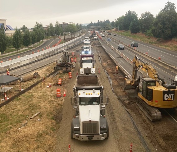 Several dump trucks lined up next to the highway with an excavator nearby