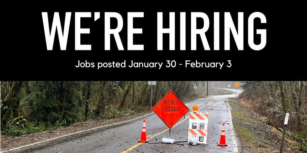 Road closed sign, two safety cones and a flashing caution light in the middle of a winding road. The surroundings appear to have some storm damage.