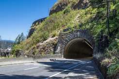 A two-lane highway tunnel in a hillside. The tunnel face is covered in masonry stone. 