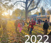 2022: children at school on the grass with fall leaves on the ground and school buses in the background