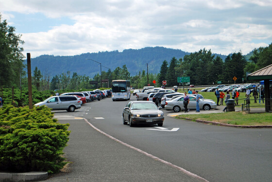 A car and tour bus circle through the parking lot. Pedestrians in the crosswalk and in the grassy area in the middle of the parking lot. 