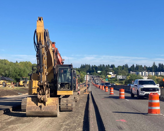 Heavy equipment in an excavated lane. Cars drive toward the camera in the left lane. The right lane is closed. 