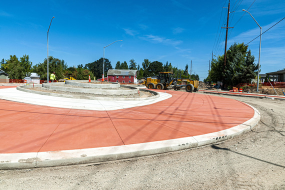 The Roundabout is full formed with salmon-colored truck aprons. The roadway is still gravel. Heavy equipment circles around the roundabout. 