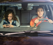Two teens in the front seat of a vehicle - one teen driving and one teen passenger texting.
