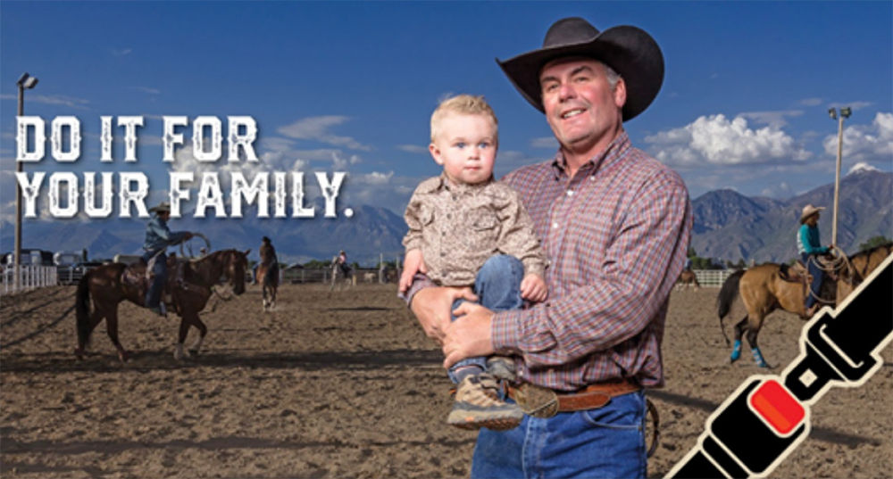 A billboard with a photo of a cowboy holding a young child. Text reads, Do it for your family. Buckle Up. Every Trip. Every Time.