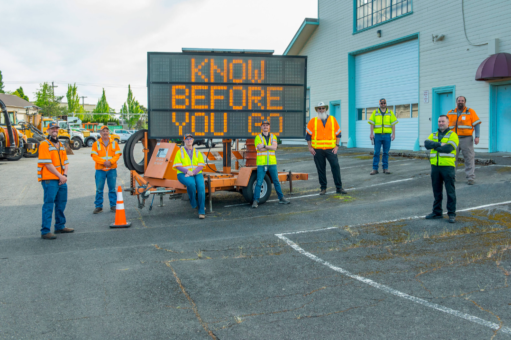 ODOT crew members stand next to a know before you go variable message road sign. 