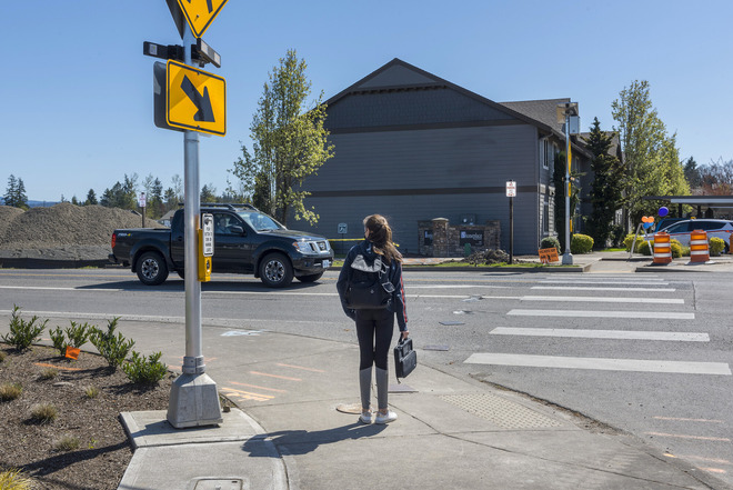 A person waits on a curb near a rapid flashing beacon pedestrian crossing, with a truck in the road.
