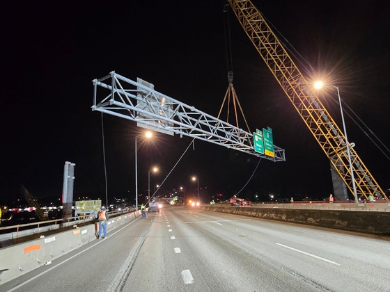 Nighttime photo of construction workers and a crane guiding an overhead highway sign structure into place.