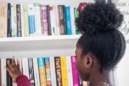Young person with dark skin and natural hair reaching toward a bookshelf 