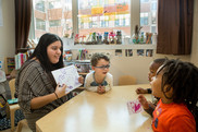 teacher sitting with young students at a table, pointing to their artwork 