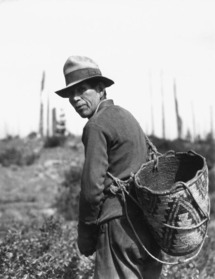 Person with a large woven basket in an outdoor setting with vegetation and tall poles in the background