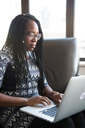Person with braided hair using a laptop near a window with buildings in the background.