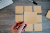 A hand holds a blank sticky note over a desk with other notes in the background.
