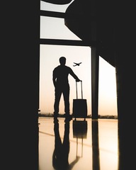 A silhouetted traveler stands at an airport window holding rolling luggage, with an airplane flying outside.