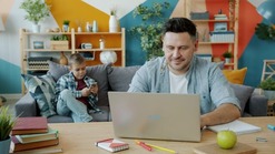 Parent sitting a a table on a laptoo with a child sitting in the background.