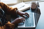 Hands typing on a laptop at a desk with a notebook and a cup of coffee nearby. 