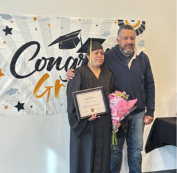 Graduate holding certificate and flowers beside another person under “Congrats Grad” banner
