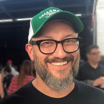Charlie Esparza smiling at camera wearing a green & white ball cap, black rimmed glasses, black shirt