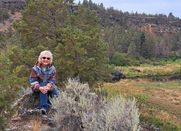 Kathy Baier wearing sun glasses, sitting on a rock surrounded by trees and brush, river flows in background