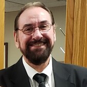 A man wearing glasses, a black suit jacket, black tie, and white shirt smiles at the camera.