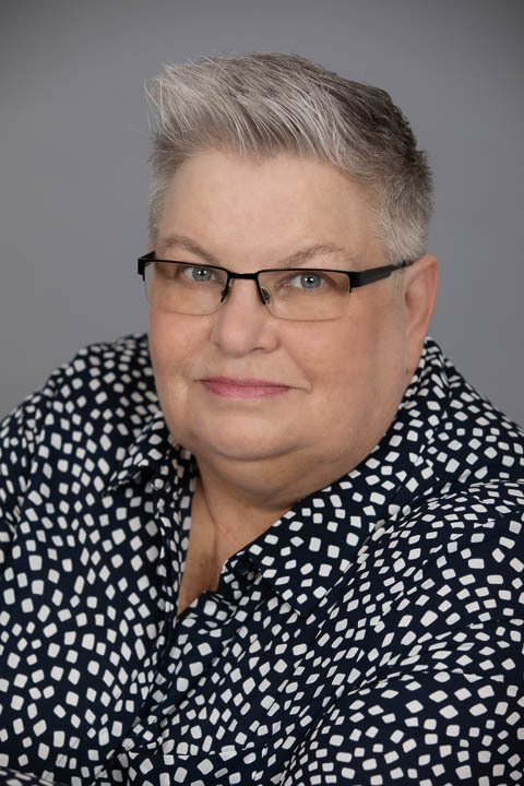 headshot of white woman with short white hair and blue eyes wearing glasses and a black and white shirt