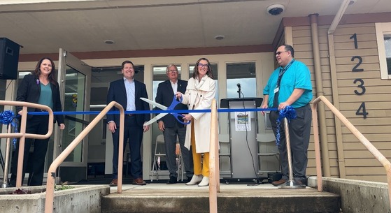 Five people standing in front of a building cutting a blue ribbon at the entrance with large scissors.