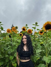 Hailey has long black hair, a nose ring and some tattoos on her arm. She is wearing a black top and skirt while standing in front of sunflowers. 